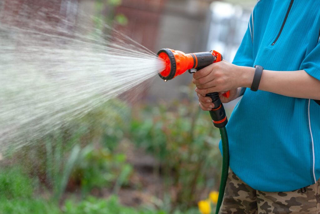 Kid/s hands watering plants from the hose, makes a rain in the garden. Gardener with watering hose and sprayer water on the flowers. Sparkling water spraying out of sprinkler on the green lawn. Summer gardening.